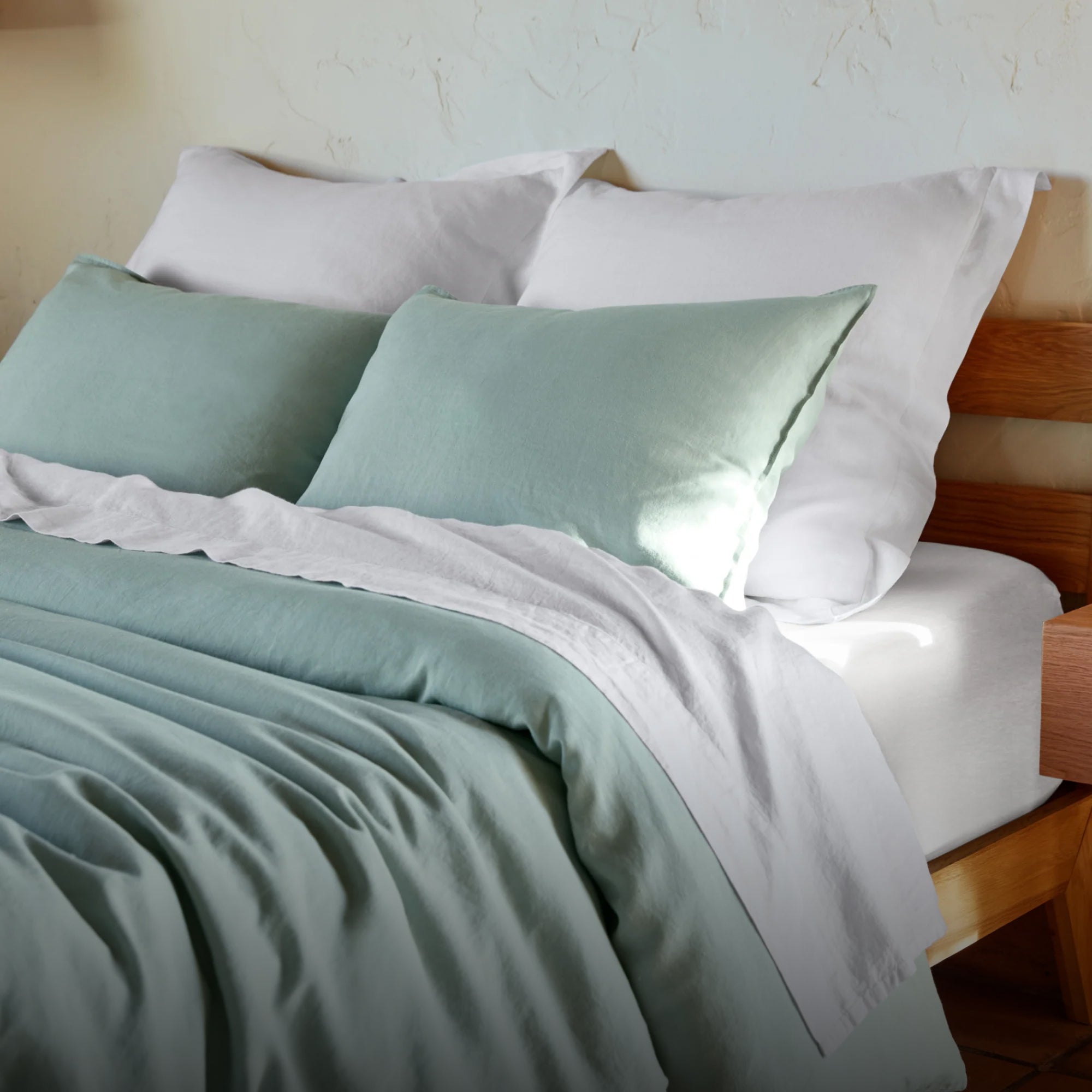 Bed made with green and white bedding and pillows in a softly lit room with a white textured wall.