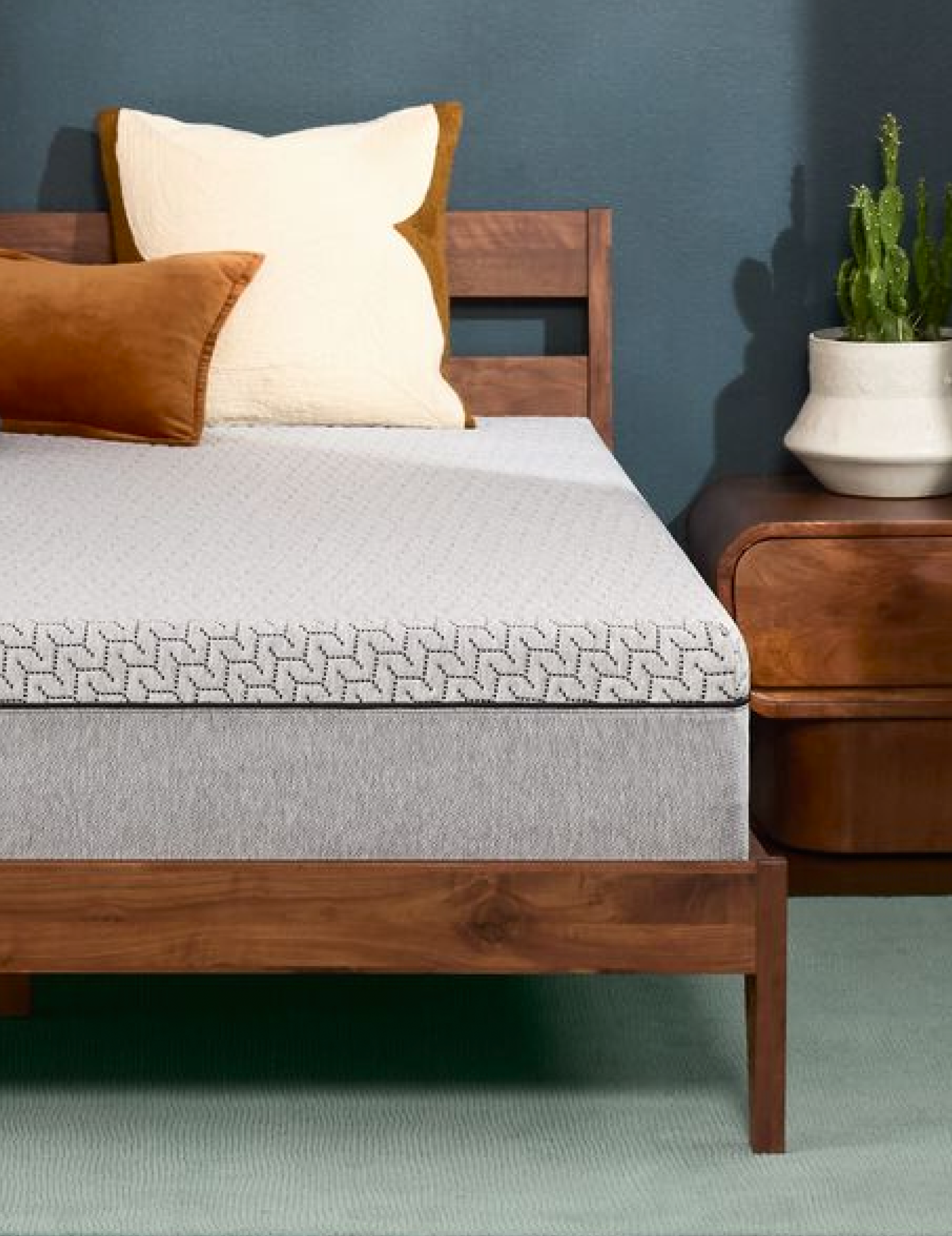 Tuft & Needle mattress on a wooden bed frame with white and rust-colored pillows beside a nightstand and small cactus.